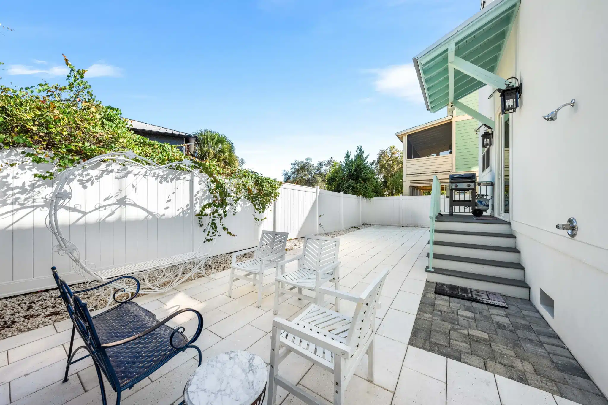 Bright backyard patio with white Adirondack chairs, pavers, and white fencing overlooking gardens